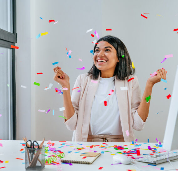Young business woman having fun time catching confetti sitting at the desk in the office. Celebrating an internship to job offer.